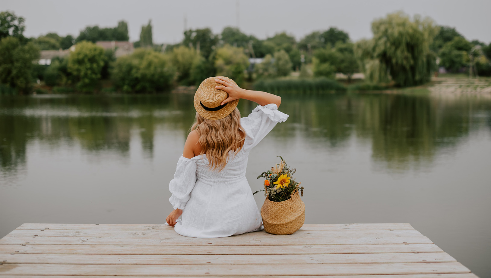 Person in white dress and straw hat with basket of flowers sitting on dock.